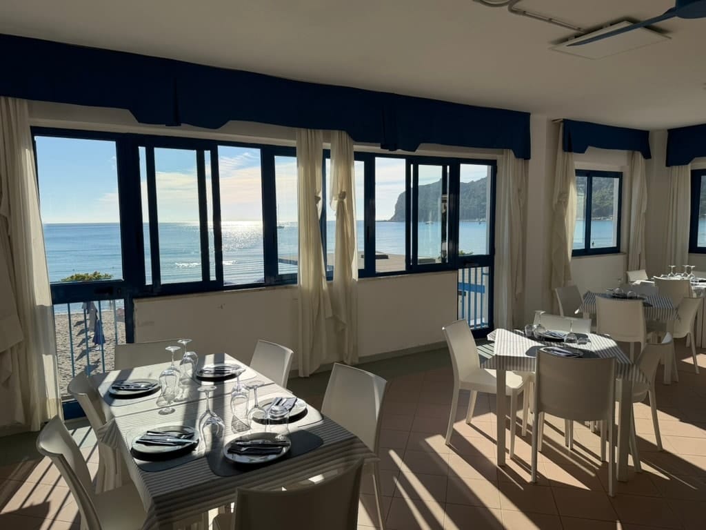 La sala al sole del mattino, con le vetrate blu sul mare e i tavoli apparecchiati